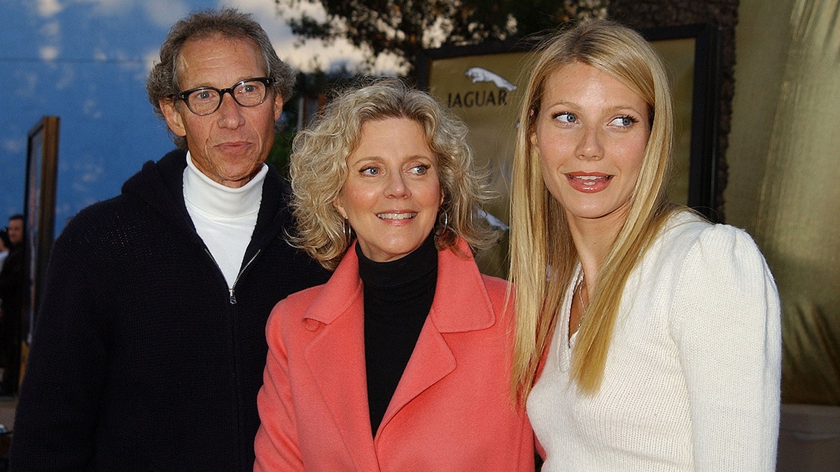 Gwyneth Paltrow in white smiles and looks to her left with mother Blythe Danner in a vibrant salmon jacket and her father Bruce in a black coat