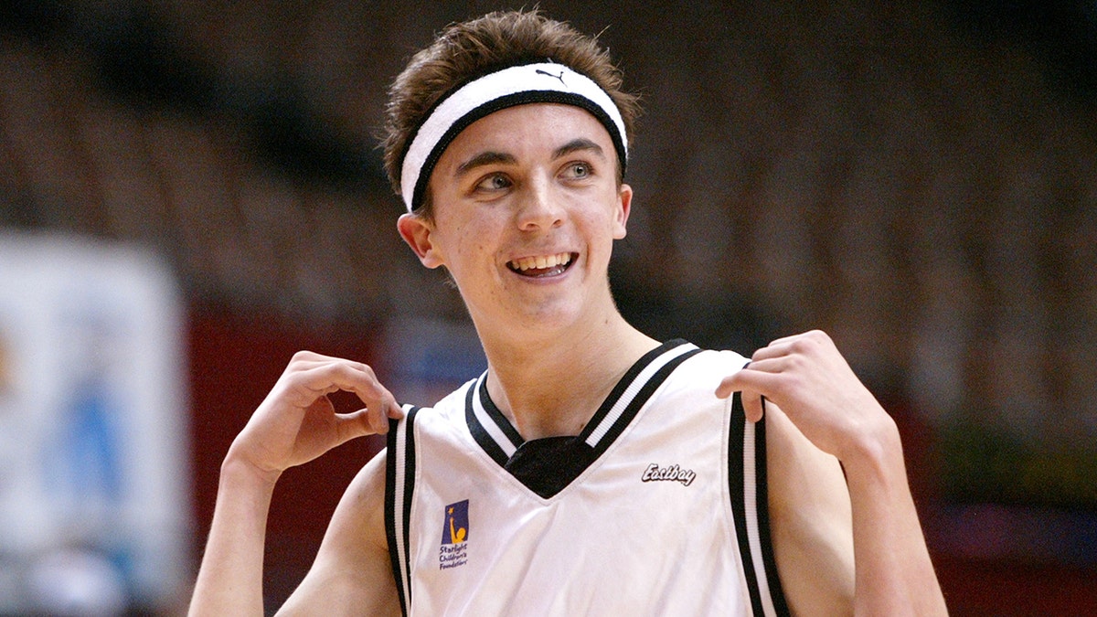 Frankie Muniz in a basketball jersey and matching sweatband holds up the sleeves of his jersey and smiles