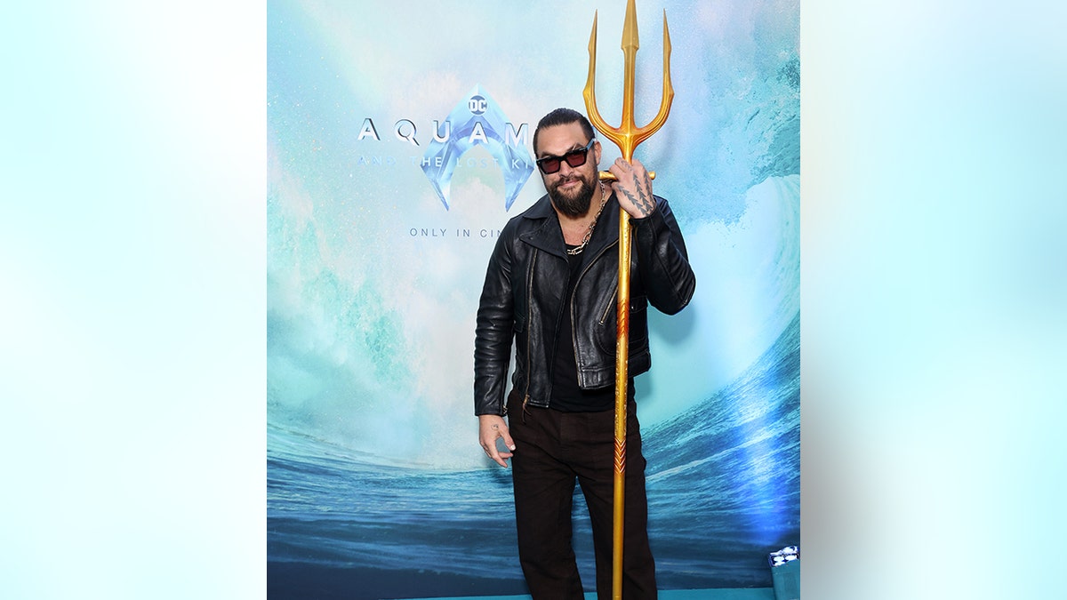 Jason Momoa in a black leather outfit holds trident on "Aquaman" red carpet