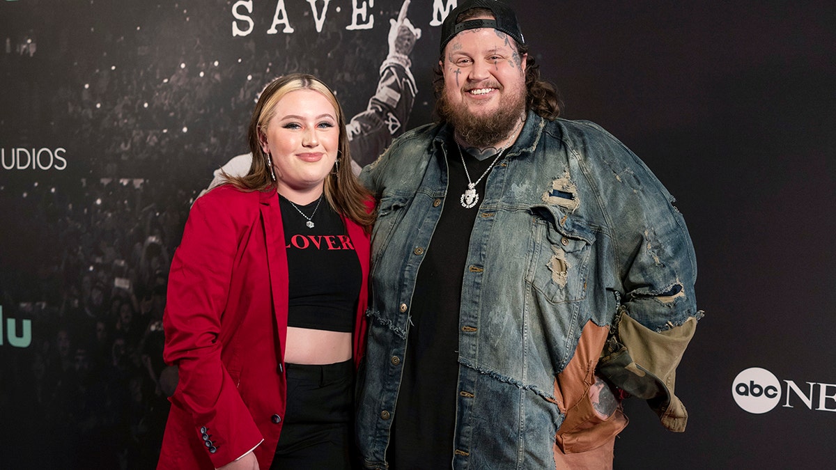 Jelly Roll in a ripped jean jacket and backward hat smiles with his daughter Bailee wearing a black crop top and red blazer