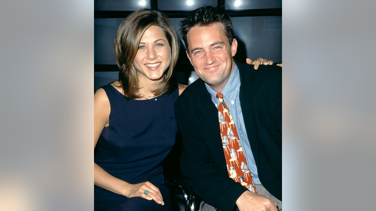 Jennifer Aniston in a navy blue dress smiles next to Matthew Perry in a black jacket, blue button-down and red patterned tie in New York