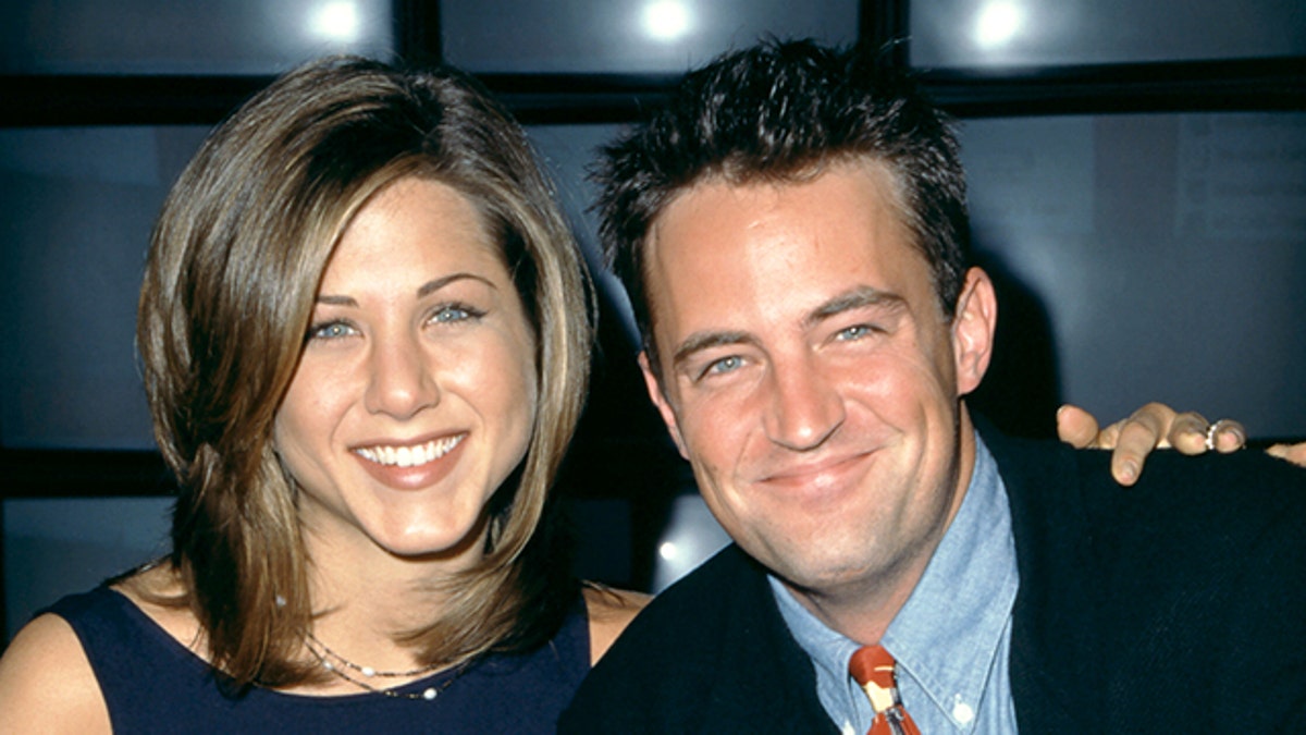 Jennifer Aniston in a navy blue dress smiles next to Matthew Perry in a black jacket and blue button down and red patterned tie in New York