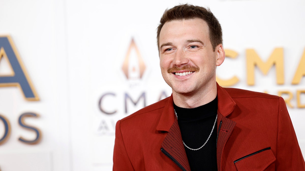 Morgan Wallen in a red suit and black t-shrit smiles on the carpet at the CMAs