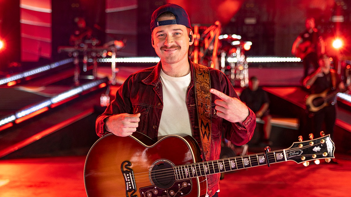 Morgan Wallen points himself on stage at the BBMAs in a cream shirt and red jean jacket