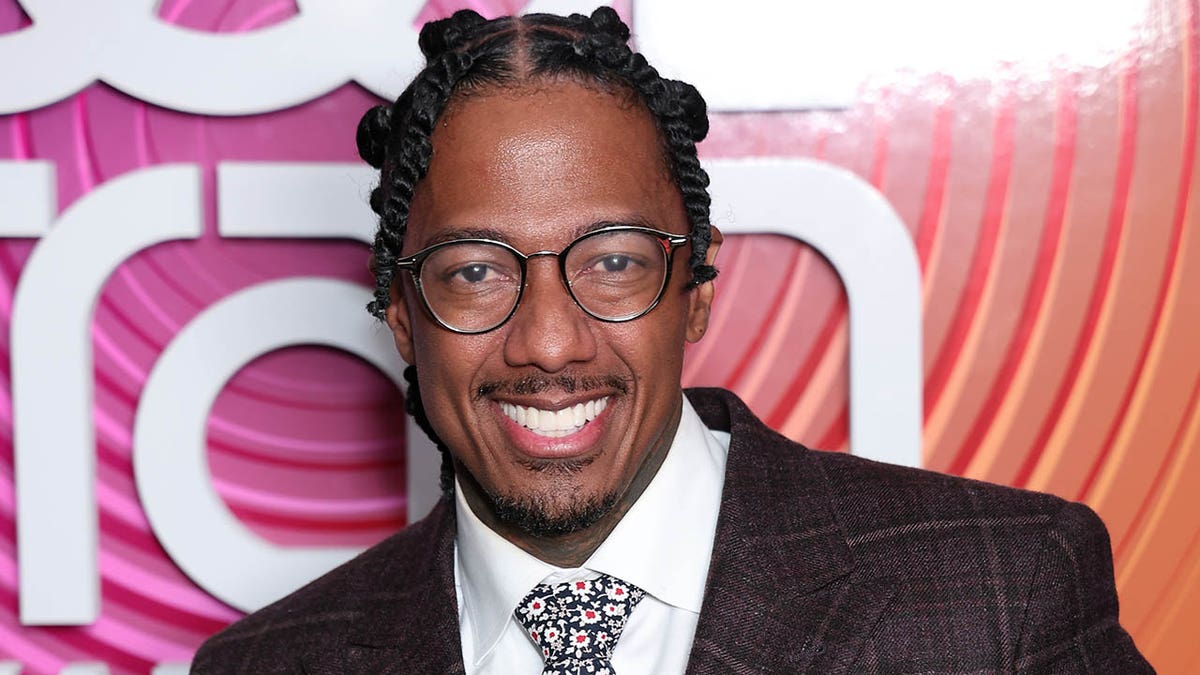 NIck Cannon in a brown suit smiles on the red carpet with circular eyeglasses
