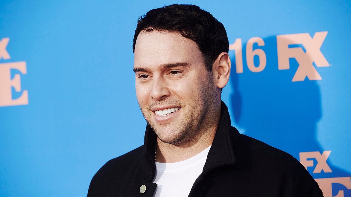 Scooter Braun in a black sweater and white shirt smiles on the carpet