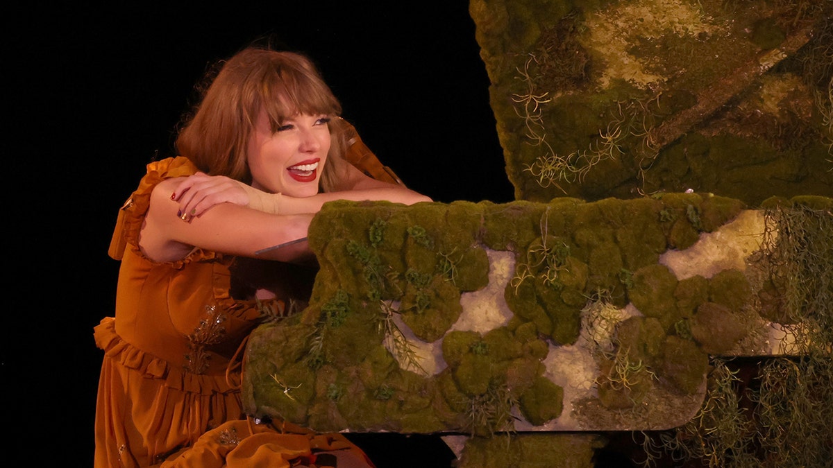 Taylor Swift smiles and lays against a moss covered piano