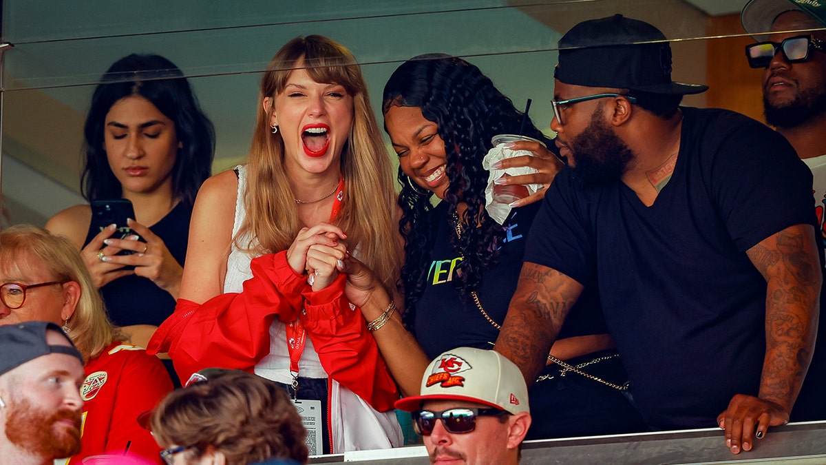 Taylor Swift in a red jacket and white tank top is surrounded by friends in a suite at the Kansas City Chiefs game