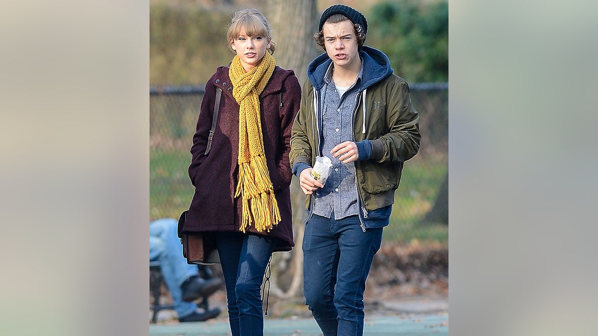 Taylor Swift in a maroon jacket and yellow scarf walks in the park with Harry Styles in a green jacket
