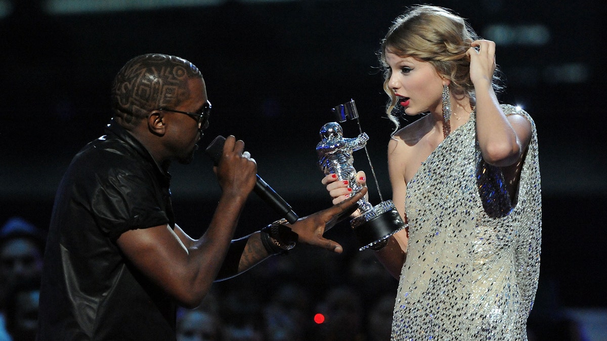 Kanye West on the VMA stage in 2009 with the microphone looks at a stunned Taylor Swift in a sparkly dress holding a moonman