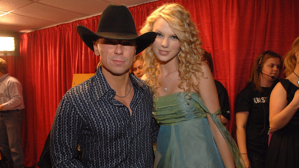 Kenny Chesney in a black cowboy hat and striped blue shirt poses with Taylor Swift in a blue and green dress