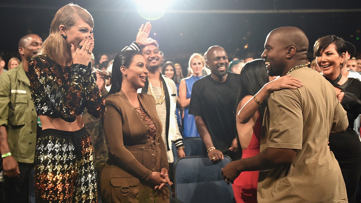 Taylor Swift covers her mouth as she looks excited towards Kanye West who looks at his wife Kim Kardashian in a brown dress