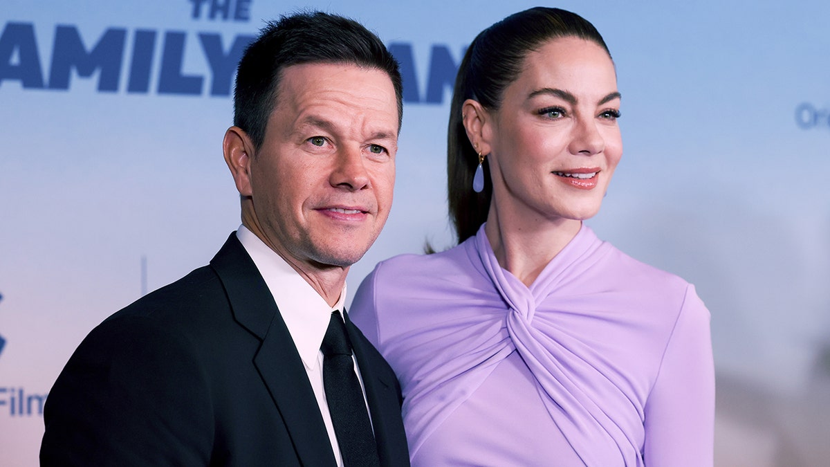 Mark Wahlberg de terno escuro e gravata está ao lado de Michelle Monaghan em um vestido lilás