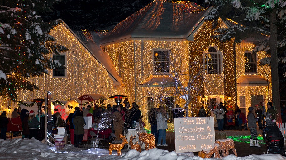 Crowd of people stand in snow outside house covered in Christmas lights