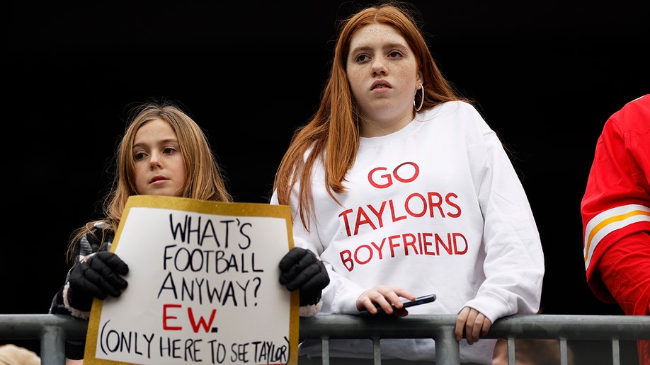 One young girl holds a sign that says "What's football anyway? Ew. Only here to see Taylor." Another girl is wearing a sweatshirt that says "Go Taylors boyfriend"