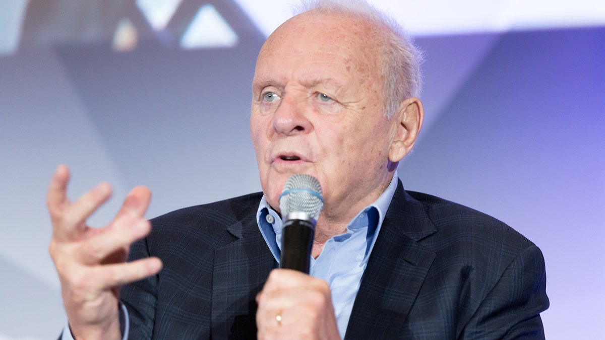 Anthony Hopkins in a dark suit and light blue shirt speaks into a microphone