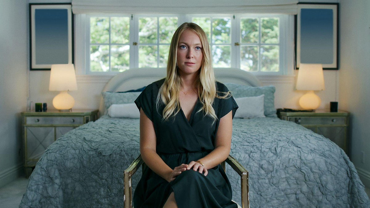 India Oxenberg sitting in front of a bed on a chair looking serious