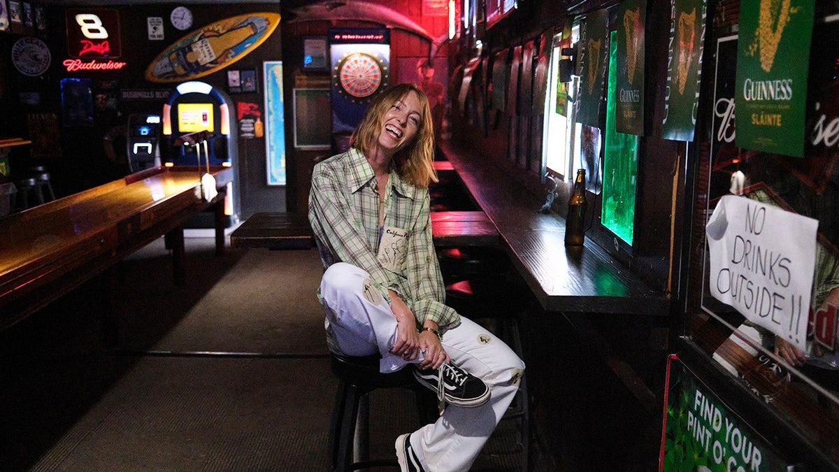 India Oxenberh wearing a plaid shirt and white pants sitting down and smiling inside a bar