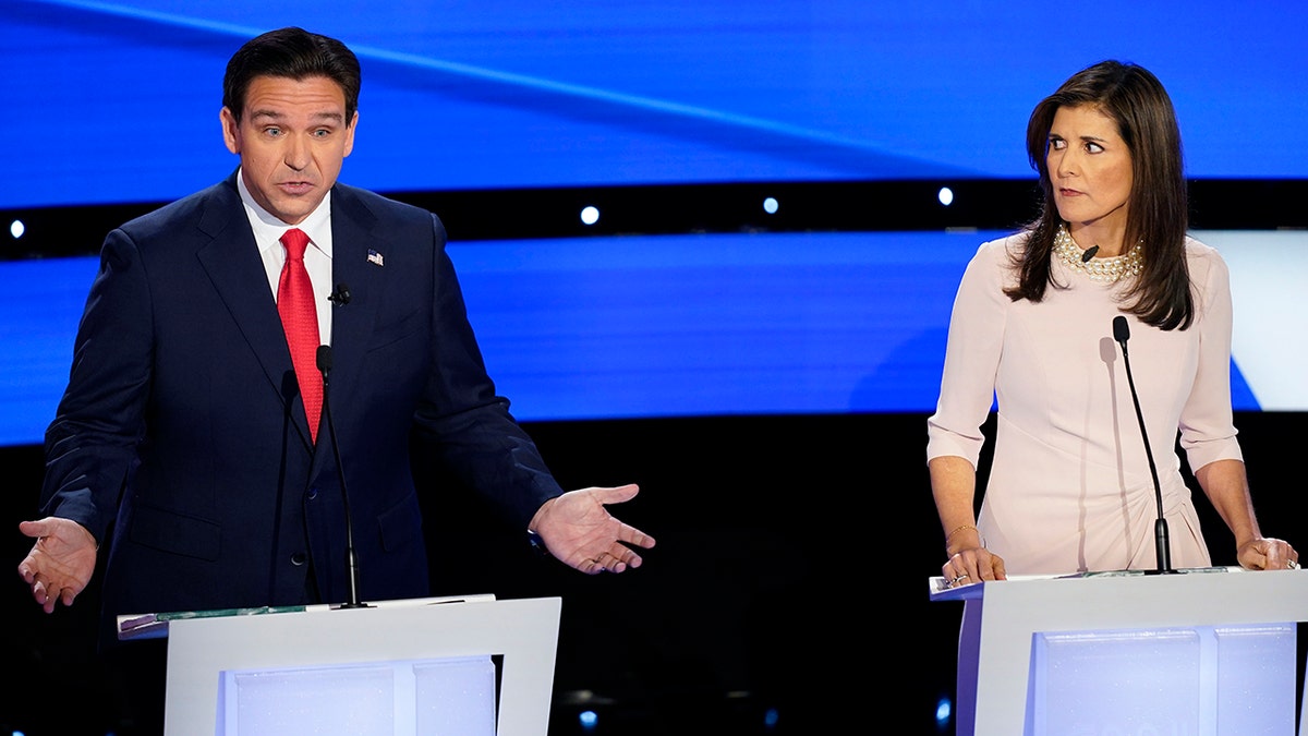 Ron DeSantis wearing navy suit, bright red tie, with arms out and hands open, talking (left), Nikki Haley wearing light pink dress, pearl necklace serious expression looking at DeSantis (right)