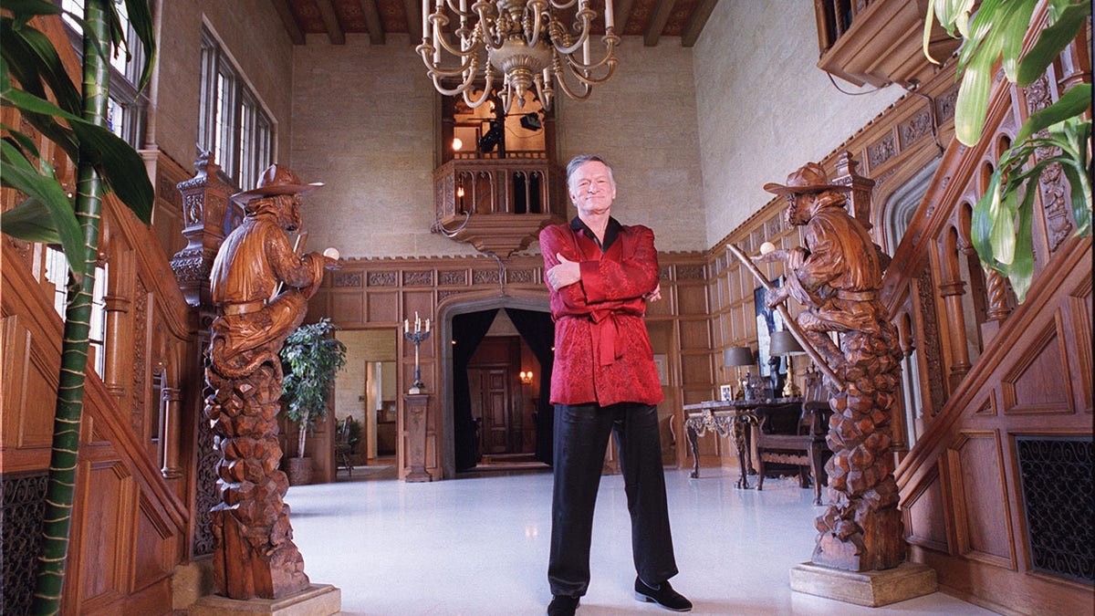 Hugh Hefner with his arms crossed wearing a red robe inside the Playboy Mansion