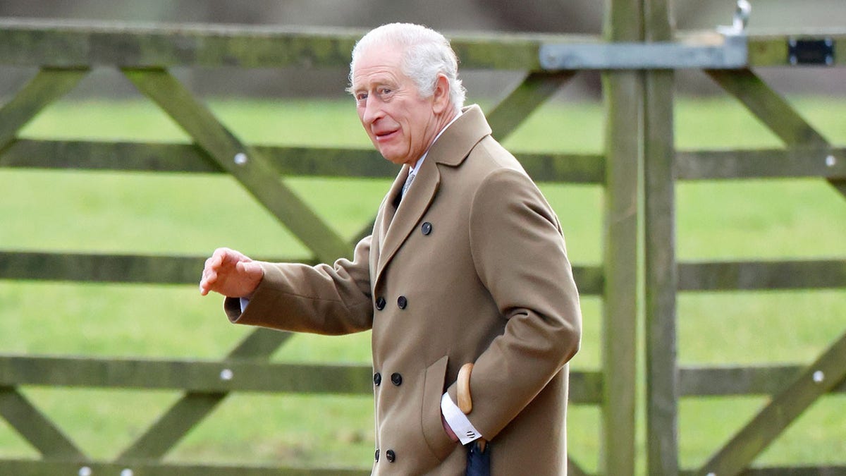 King Charles wearing a brown coat and walking next to a fence