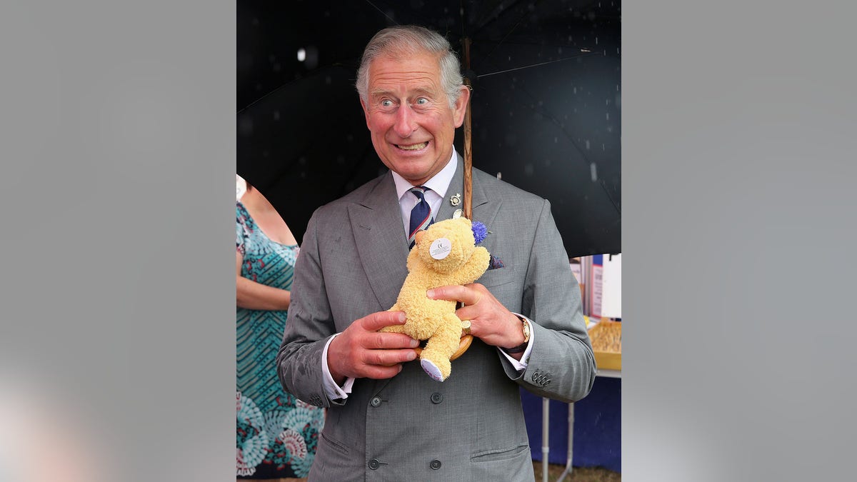 King Charles smiling and holding a yellow teddy bear and an umbrella