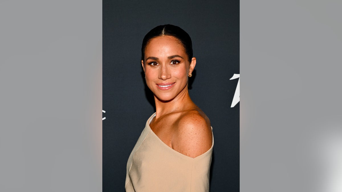 Meghan Markle standing to the side and showing off her shoulder in a beige dress