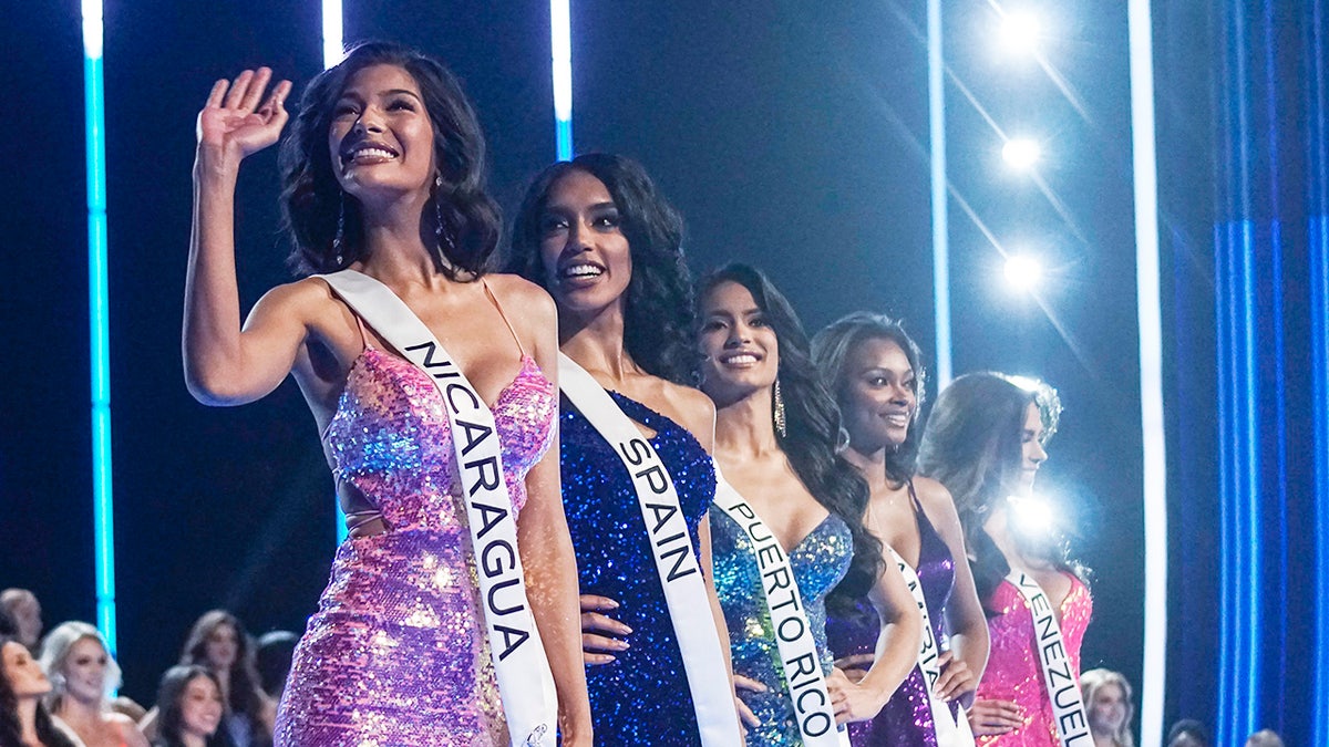 A group of Miss Universe contestants wearing sashes and gowns waving to the side