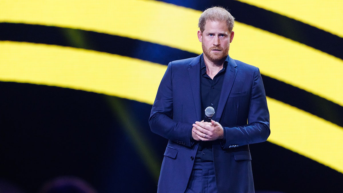 Prince Harry wearing a blue suit and black shirt holding a mic