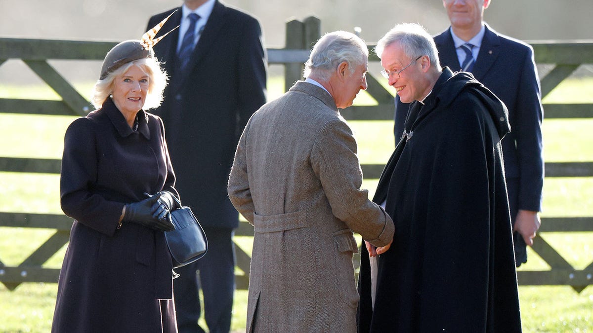 Queen Camilla holding onto a purse as King Charles shakes hands with a priest