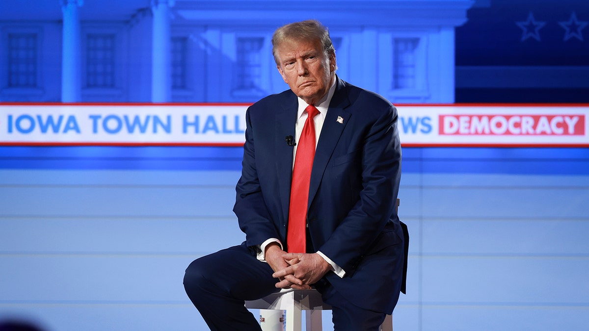 Trump con semblante serio, sentado en un taburete del escenario, con las manos cruzadas sobre el regazo, vestido con traje azul marino y corbata roja brillante