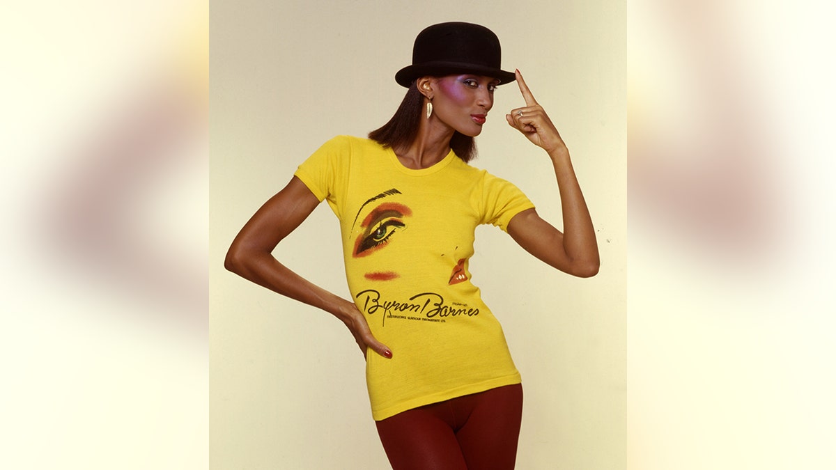 Beverly Johnson in a bright yellow patterned dress with a hat and one hand on her hip