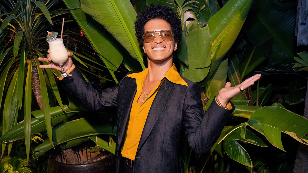 Bruno Mars com camisa amarelo mostarda com gola longa e terno escuro colocando as mãos para o lado segurando uma bebida