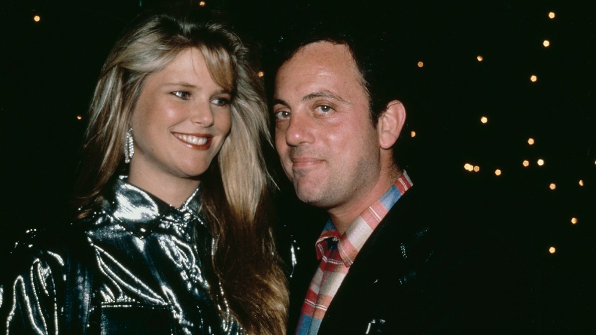 Christie Brinkley sorri e olha levemente para Billy Joel em uma jaqueta preta e camisa de colarinho estampada