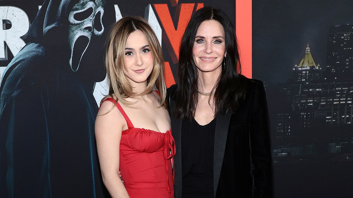 Coco Arquette in a red dress poses with her mother Courteney Cox in a black velvet jacket on the carpet