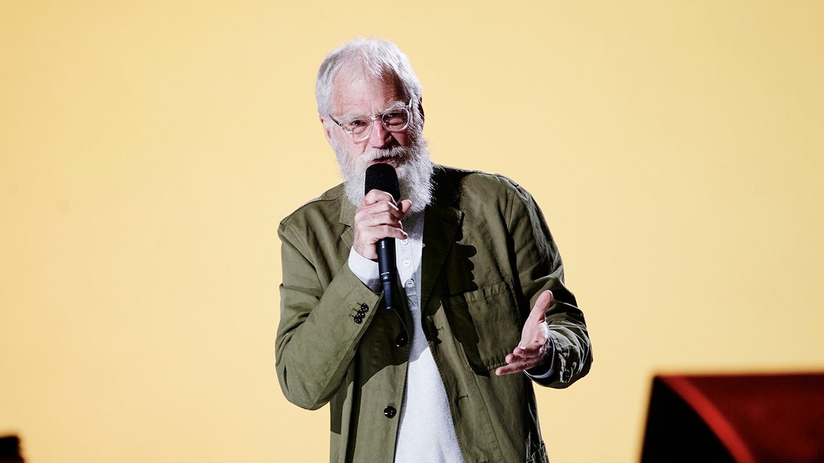 David Letterman in an olive green jacket speaks with his hands on stage