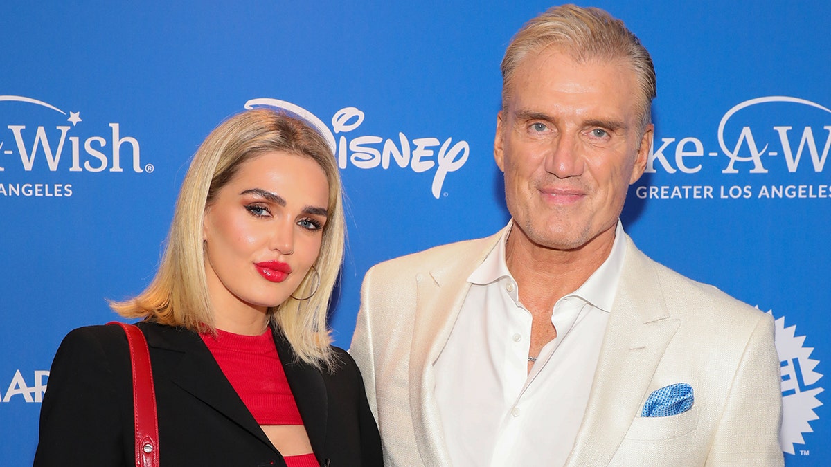 Emma Krokdal in a black blazer and red shirt poses on the carpet with Dolph Lundgren in a cream blazer and white shirt
