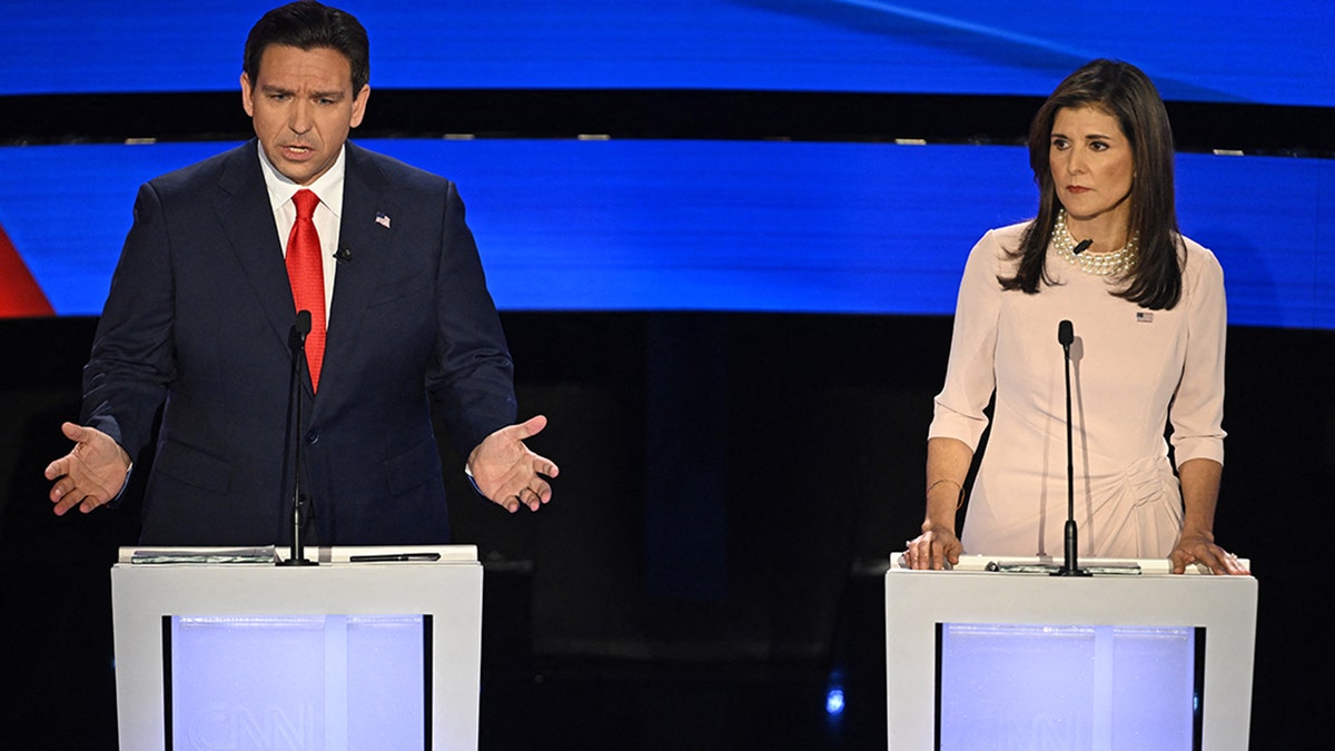 Ron DeSantis wearing navy suit, bright red tie, with arms out and hands open, talking (left), Nikki Haley wearing light pink dress, pearl necklace serious expression (right)