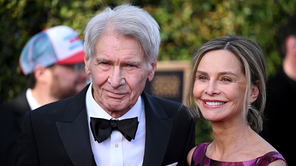 Harrison Ford in a classic tuxedo smiles at the Critics Choice Awards with wife Calista Flockhart in purple