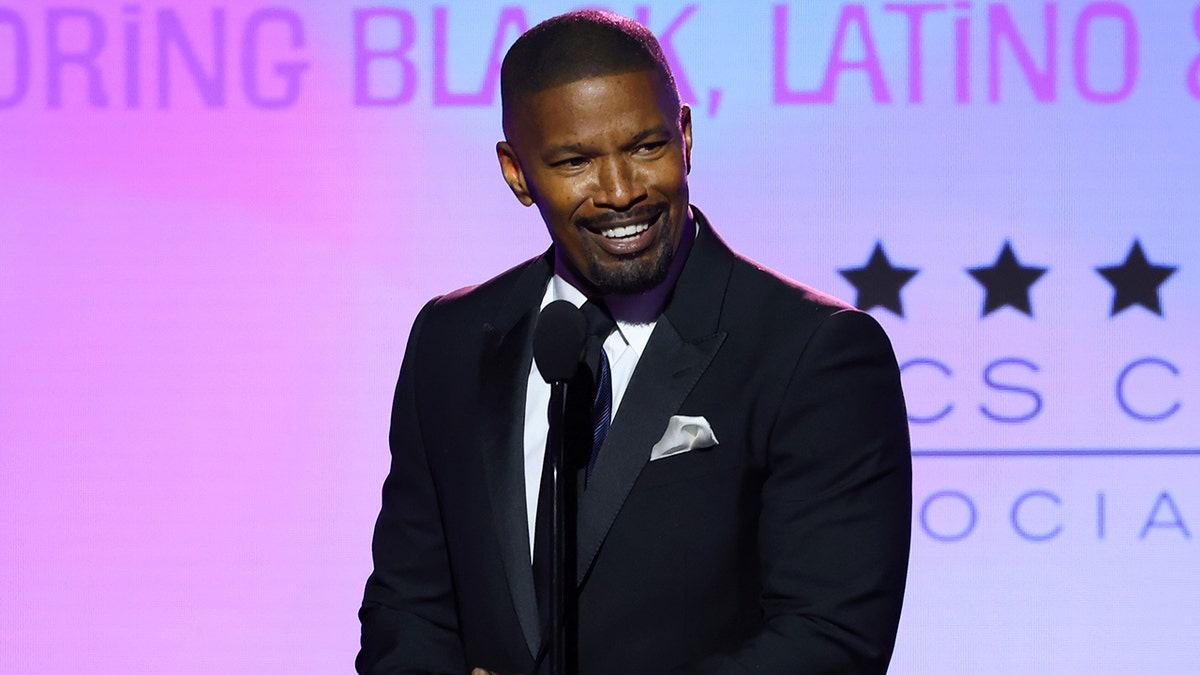 Jamie Foxx in a black suit smiles and stands in front of a microphone on stage