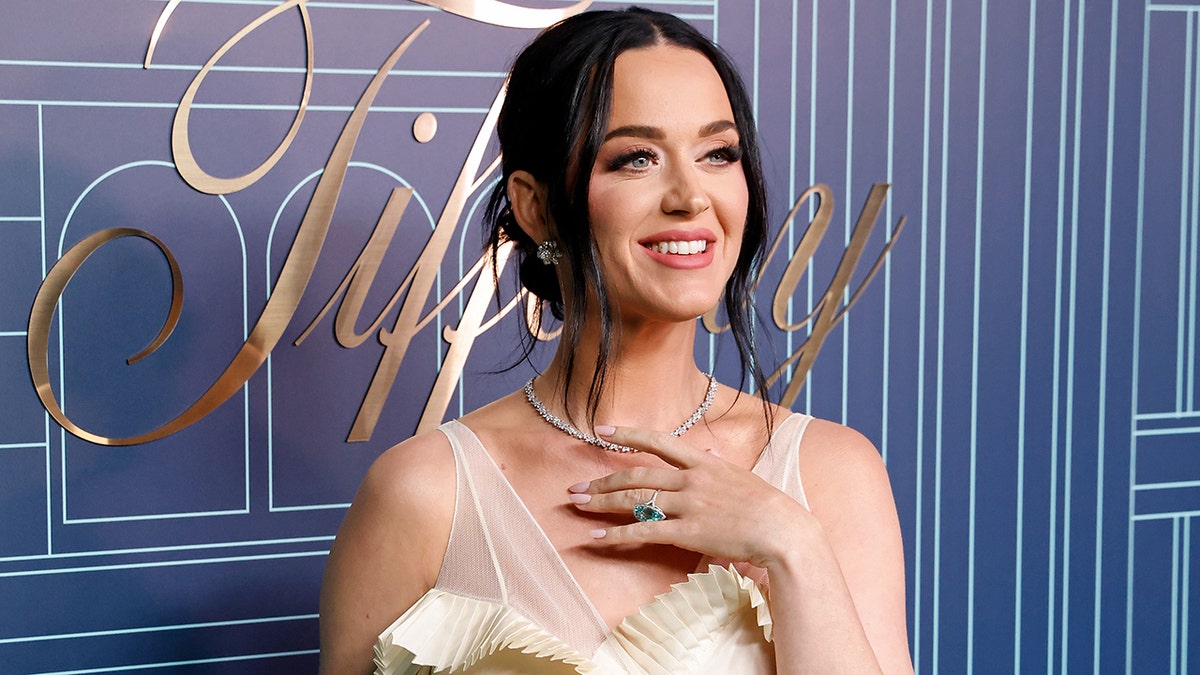 Katy Perry with her hand on her chest looks away from the camera in a champagne colored dress