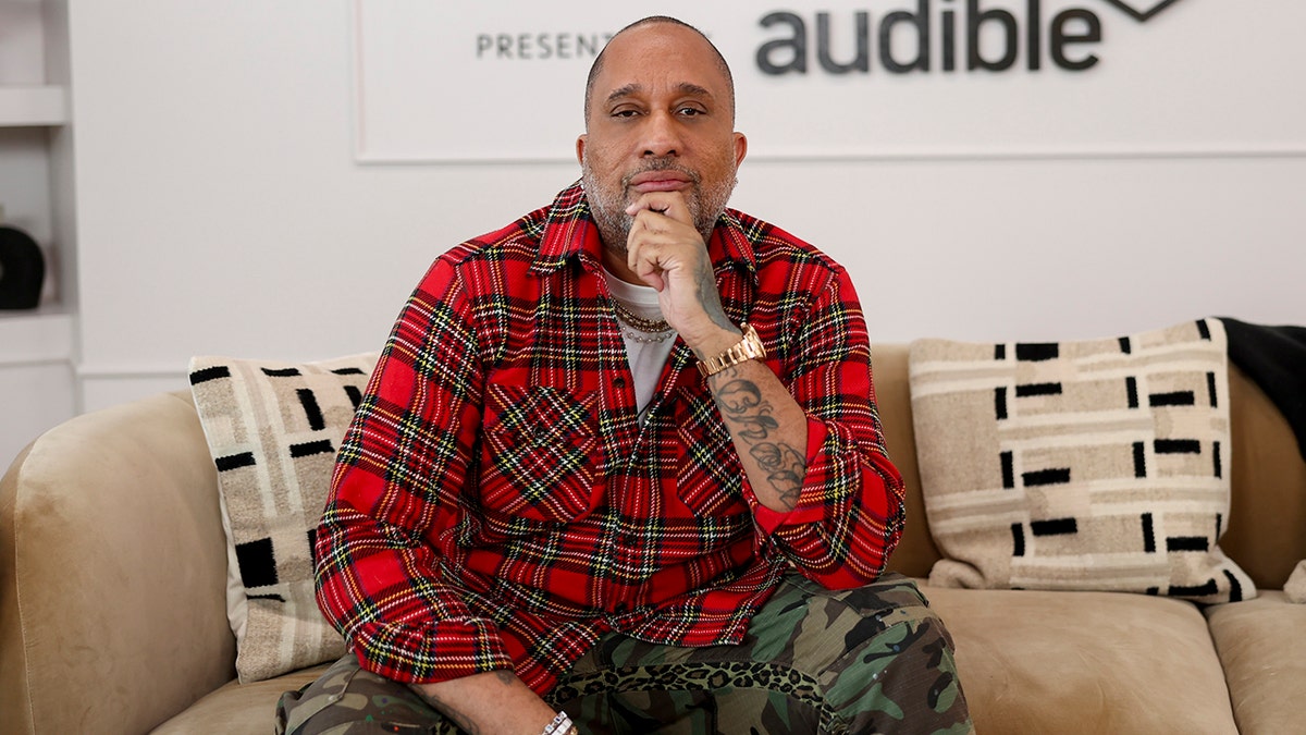 Kenya Barris a red plaid shirt puts his hand to his chin and sits on a couch