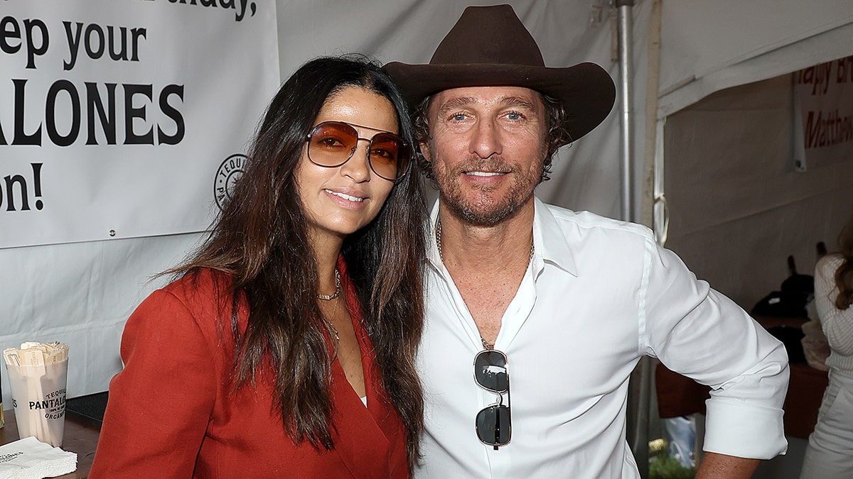 Matthew McConaughey in a white shirt and cowboy hat smiles with wife Camila Alves in a red top