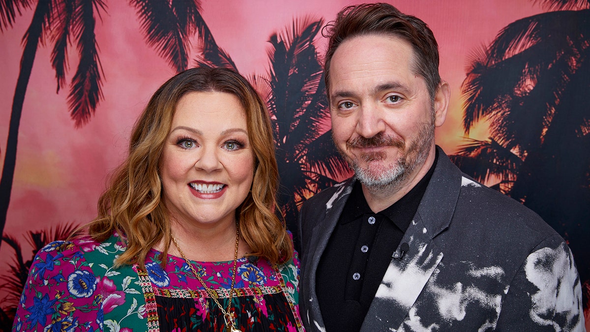 Melissa McCarthy in a patterned dress smiles alongside Ben Falcone, soft smiling