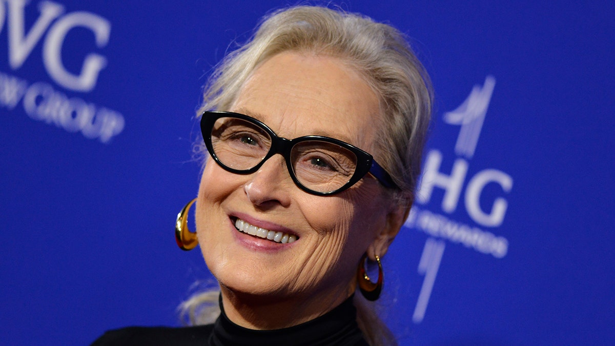 Meryl Streep smiles wearing large gold earrings and black cat eye glasses