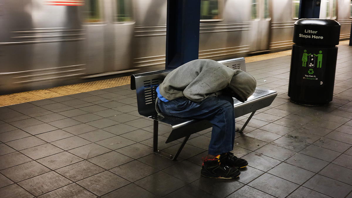 Homeless man asleep on subway station bench, train passes behind him