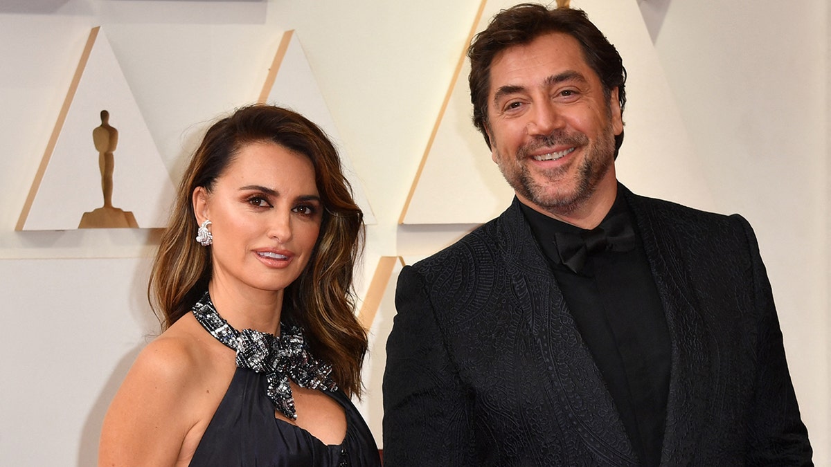 Penélope Cruz in a navy halter gown poses on the carpet with husband Javier Bardem in a black shirt and suit jacket