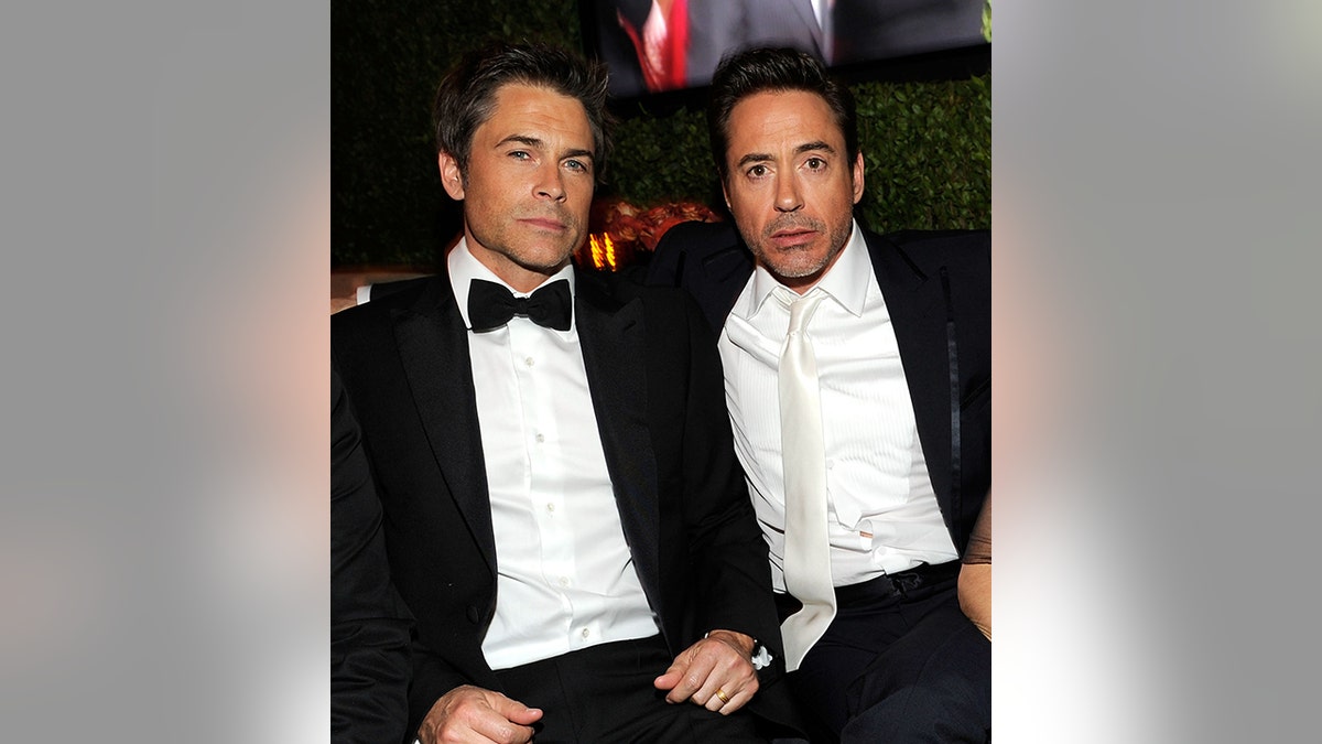 Rob Lowe in a classic tuxedo sits next to Robert Downey Jr. in black suit and white shirt and tie