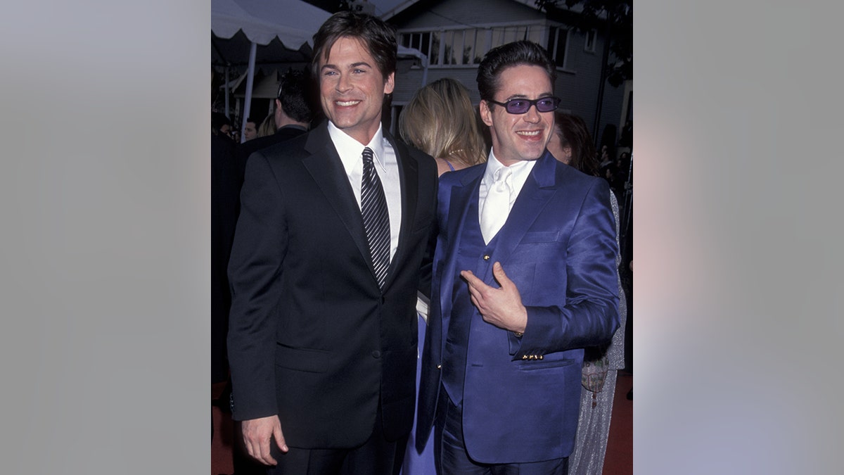 Rob Lowe de terno preto sorri e olha para a direita enquanto Robert Downey Jr. aponta para ele de terno azul e olha para a esquerda