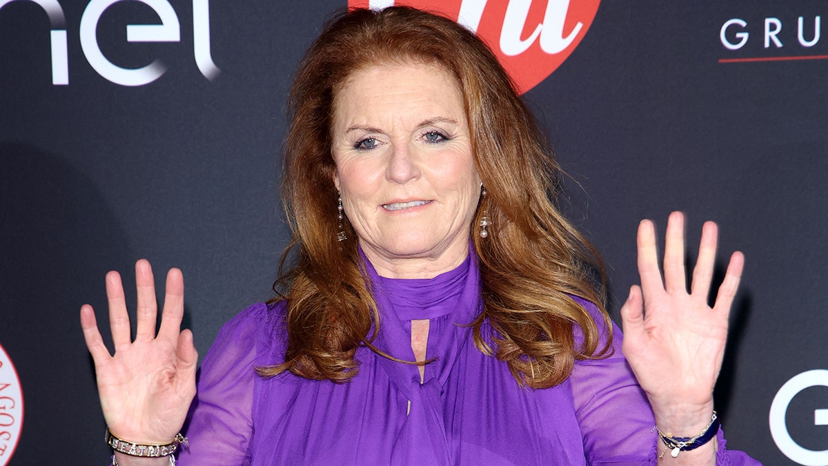 Sarah Ferguson in a purple gown waves her hands on both sides on the carpet